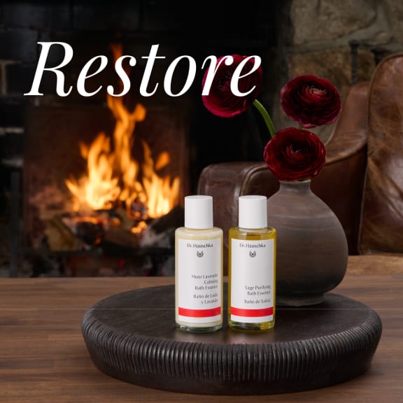 Two Dr. Hauschka bath essence bottles on a dark tray, with red flowers and a fireplace behind the word “Restore”.