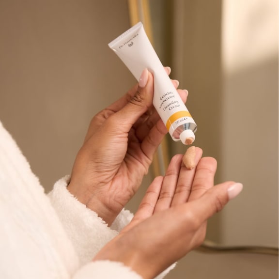 Hands squeeze cleansing cream from a tube into a palm, against a warm beige bathroom background.
