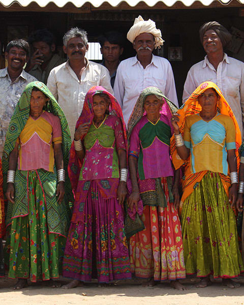 A group of ten adults stand together outside a building, with four women in the front wearing brightly colored traditional clothing and head coverings, and six men standing behind them.