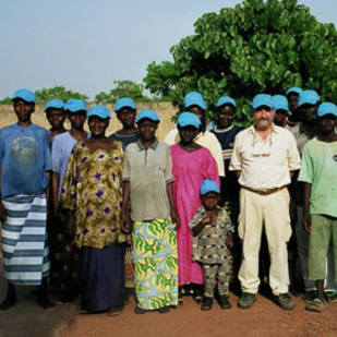 Group of people standing outdoors on red earth, many wearing blue caps, with one tree and a building in the background.