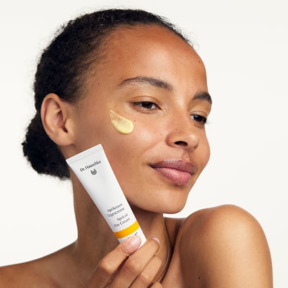 Woman applies cream to her cheek while holding a Dr. Hauschka Apricot Day Cream tube.