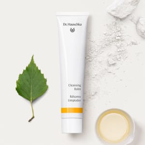 Dr. Hauschka Cleansing Balm tube beside a green leaf, white powder and a small dish of balm on a white surface.