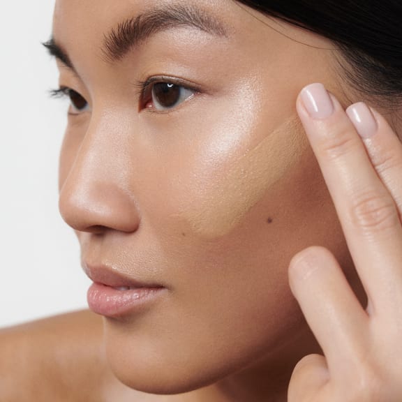 Close-up of a woman applying beige foundation to her cheek with her fingers.