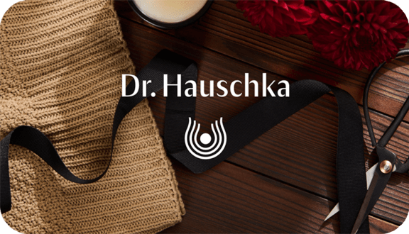 Dr. Hauschka logo on a wooden table with woven fabric, black ribbon, flowers and scissors.