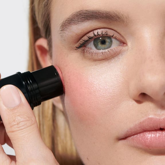 Close-up of a woman applying pink makeup blush to her cheek with a black stick applicator.