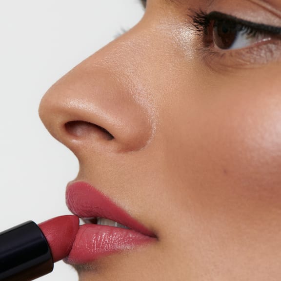 Close-up of a woman applying red lipstick to her lips, with natural makeup and a soft white background.