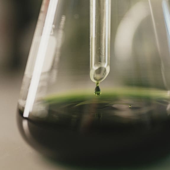 Close-up of a glass pipette releasing a green droplet into a lab flask.
