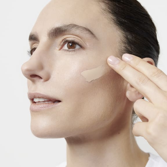 Close-up of a woman applying foundation to her cheek with her fingers, showing a makeup swatch.