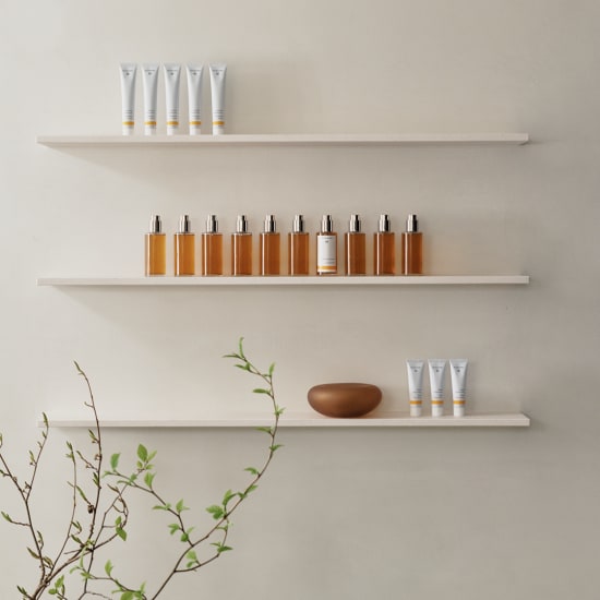 Minimal spa display with skincare tubes, amber bottles on floating shelves, a brown bowl, and plant branches.