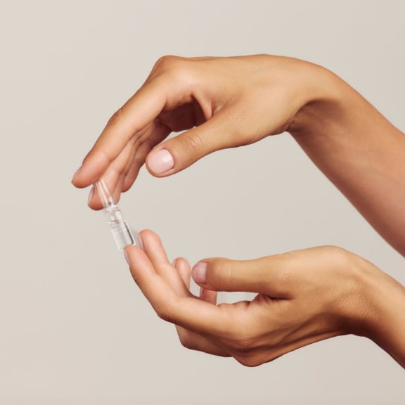 Two hands holding a small clear ampoule against a plain beige background.