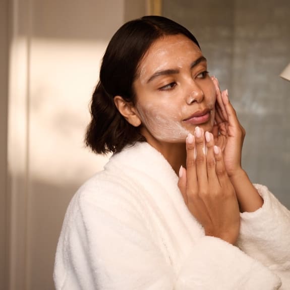 Woman in a white robe applying facial cleanser in a bathroom, with cream on her cheek and chin.