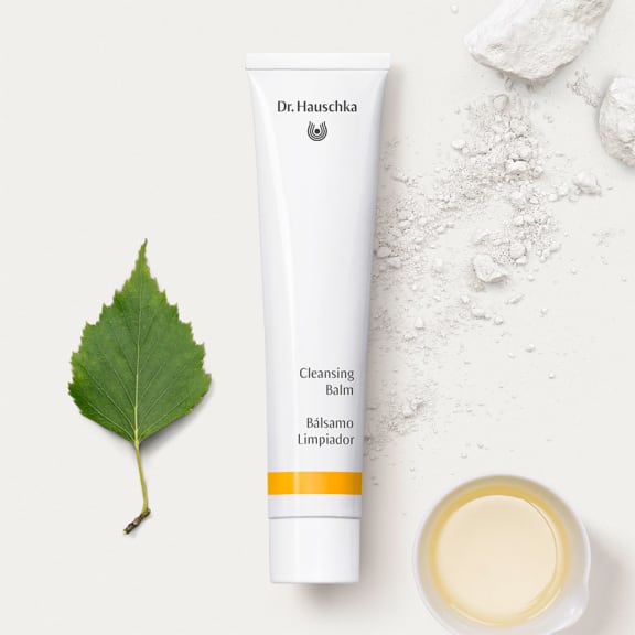 Dr. Hauschka Cleansing Balm tube beside a green leaf, white powder and a small dish of balm on a white surface.
