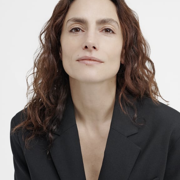 Portrait of a woman with wavy brown hair in a black blazer against a white background.