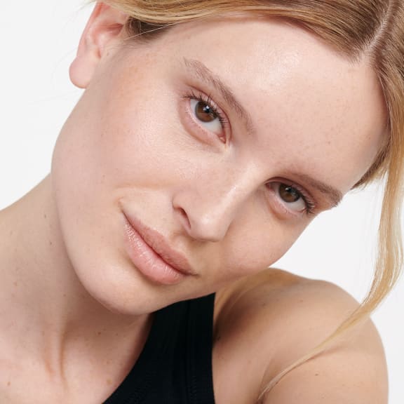 Close-up portrait of a woman with natural makeup, freckles and tied-back blonde hair against a white background.