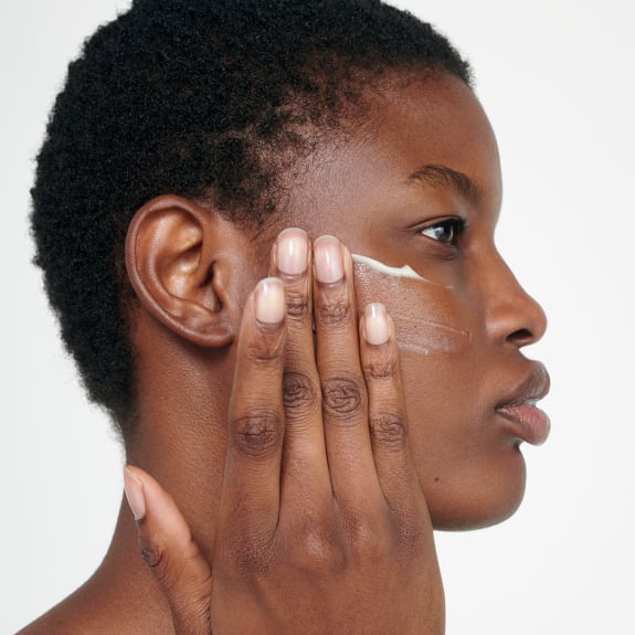 Side profile of a woman applying lotion under her eye with her fingers against a white background.