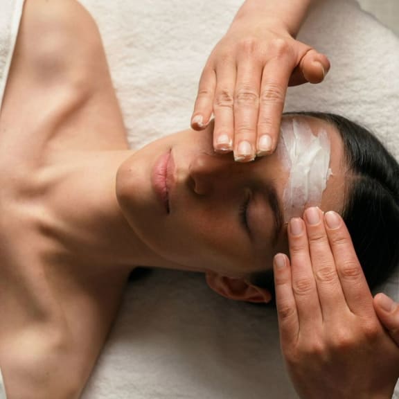 Woman receiving a facial mask treatment, with hands applying cream to her forehead in a spa setting.