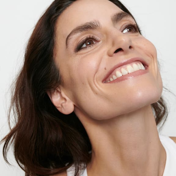 Close-up of a smiling woman with natural makeup, looking upward against a white background.