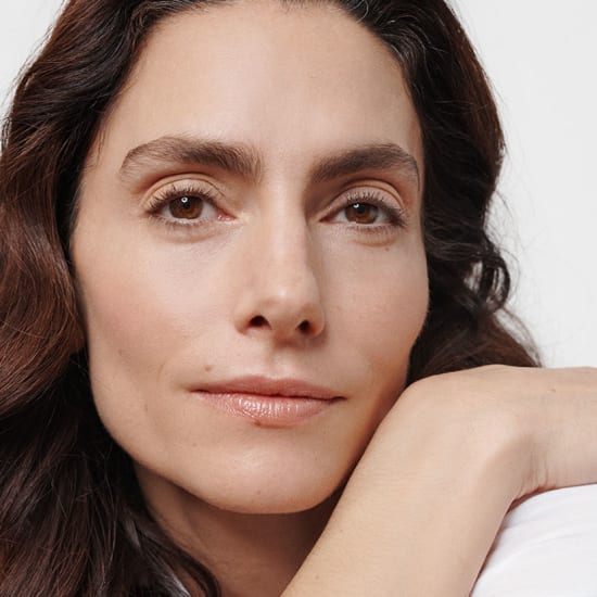 Close-up portrait of a woman with natural makeup resting her chin on her hand against a white background.