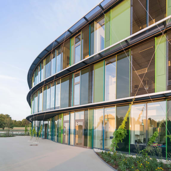 Modern curved office building with glass facade, green panels and landscaped entrance under a clear sky.