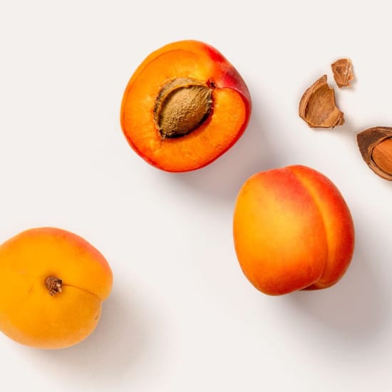 Three apricots on a white background, one halved with the pit visible and shell pieces nearby.