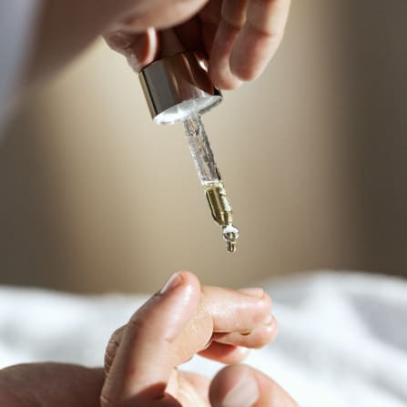 Close-up of a dropper dispensing serum onto a fingertip, with a blurred hand holding it above.