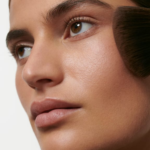 Close-up of a model’s face with soft makeup and a brush near the cheek.