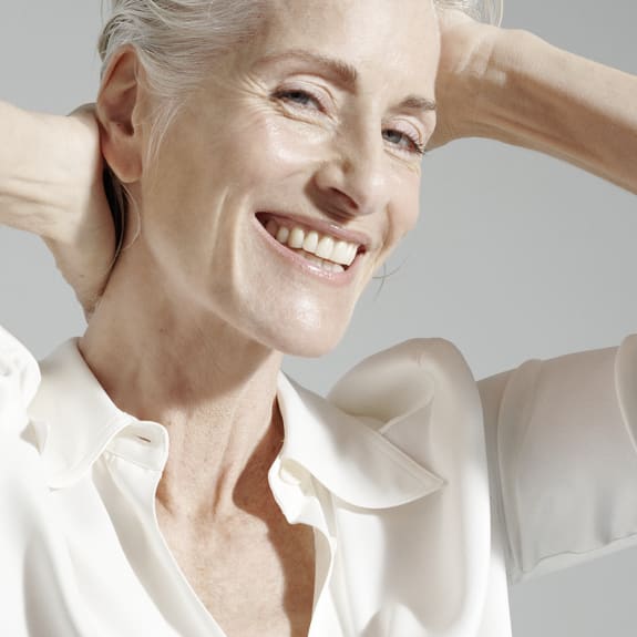 Smiling older woman in a white blouse, hands behind her head, lit by soft light against a grey background.
