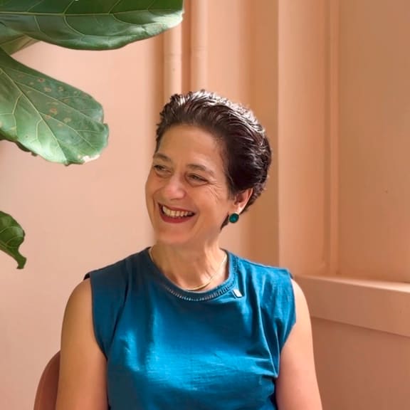 Smiling woman in a teal sleeveless top sits indoors beside large green leaves and a peach wall.