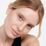Close-up portrait of a woman with freckles and tied-back blonde hair, tilting her head against a white background.