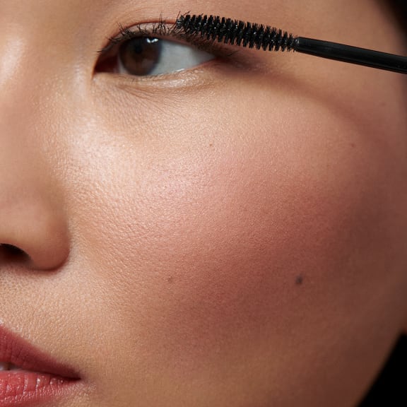 Close-up of a person applying mascara to curled eyelashes, showing glowing skin and glossy lips.
