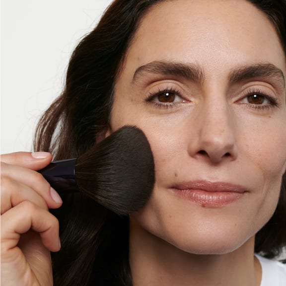 Close-up of a woman applying blush with a large makeup brush to her cheek.