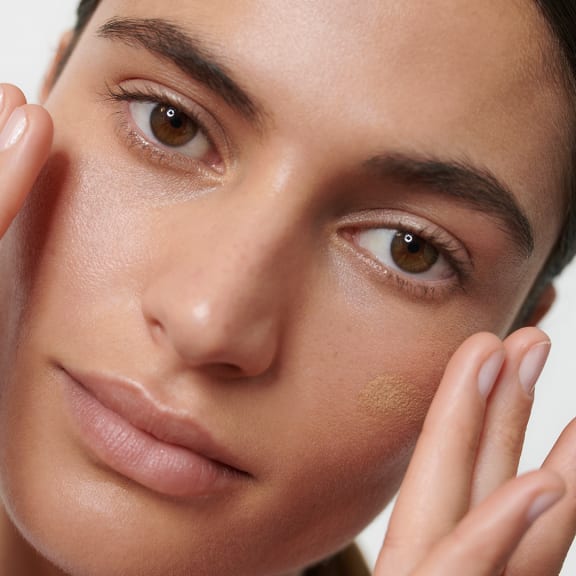 Close-up of a woman applying nude makeup, with a dab of foundation on her cheek and her fingers near her face.
