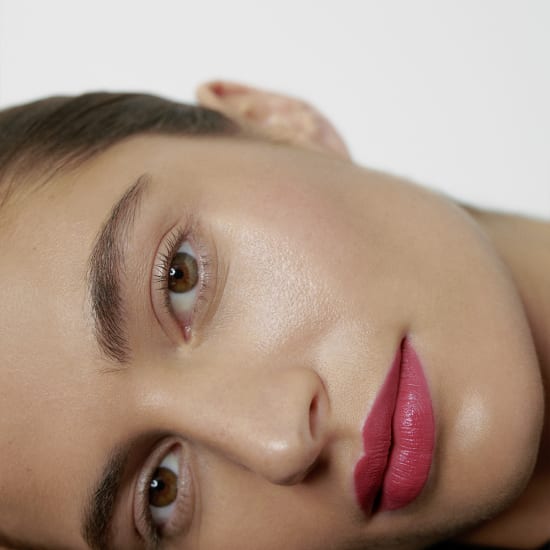 Close-up of a woman’s face with bright pink lipstick and natural makeup, shot sideways against a plain white background.