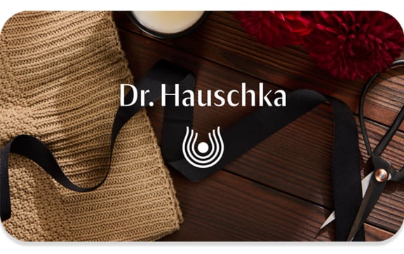 Dr. Hauschka gift card with black ribbon on woven mat, scissors, candle and red flowers on a wooden table.