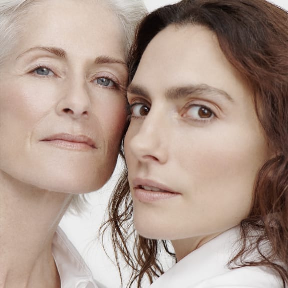 Close-up portrait of two women in white shirts, one older with gray hair and one younger with dark hair, looking at the camera.