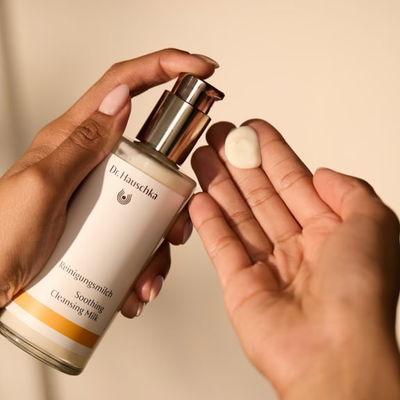 Hands holding a Dr. Hauschka soothing cleansing milk bottle, with a dollop of lotion on one palm.