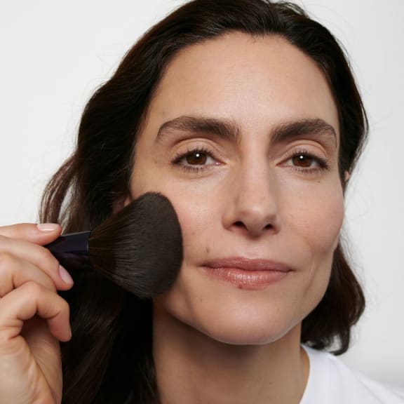 Close-up of a woman applying powder to her cheek with a makeup brush.
