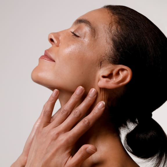 Side profile of a woman with eyes closed, gently touching her neck and jawline against a plain background.