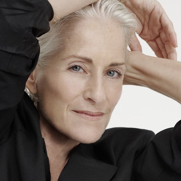Close-up portrait of a silver-haired woman in a black blazer, hands raised above her head against a white background.