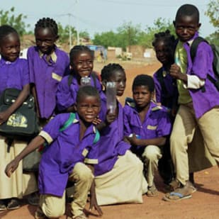 Group of schoolchildren in purple uniforms posing outdoors on a dusty road in a rural village.