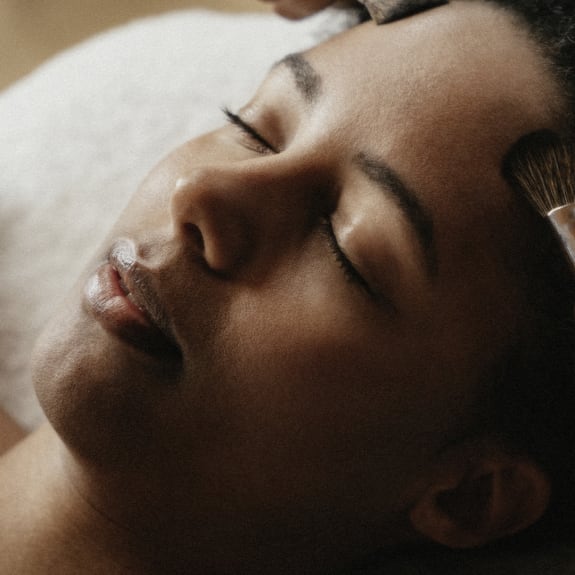 Close-up of a woman with eyes closed while makeup is gently applied with a brush.