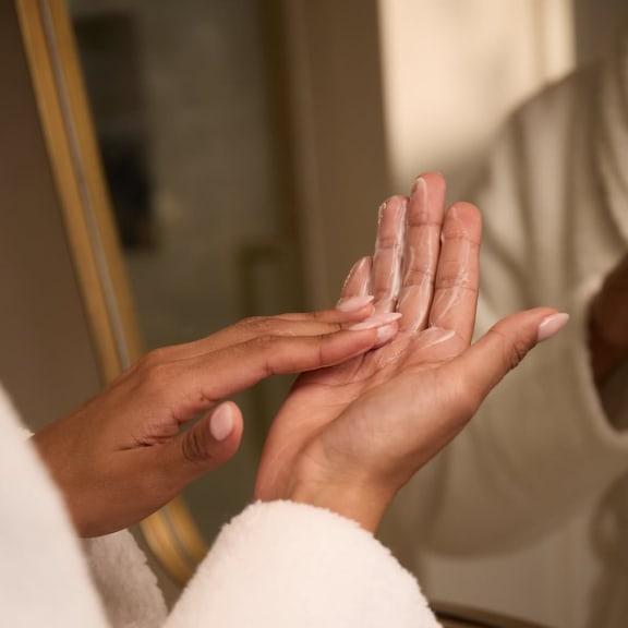 Hands applying cleansing cream to a palm in front of a mirror.
