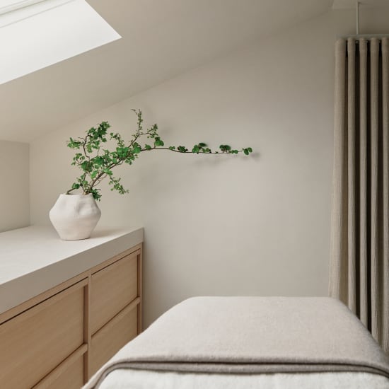 Minimal spa room with treatment bed, wooden cabinet, white vase and green branch on a pale wall.