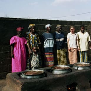 Five women and one man stand by large pots on an outdoor cookfire against a dark wall.