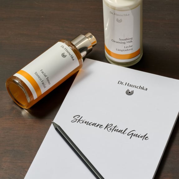 Dr. Hauschka skincare ritual guide beside a facial toner, cleansing milk and pencil on a wooden table.