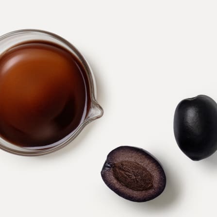 Top view of a bowl of dark sauce beside two black olives, one whole and one halved, on a white background.