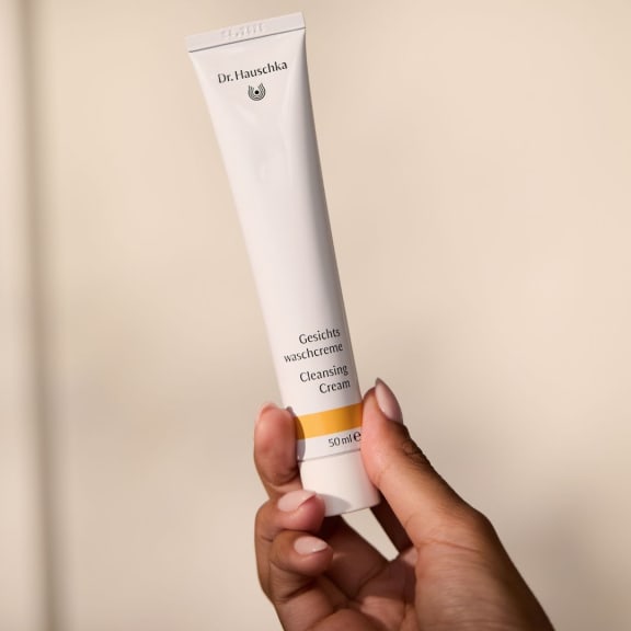 Hand holding a Dr. Hauschka Cleansing Cream tube against a plain beige background.
